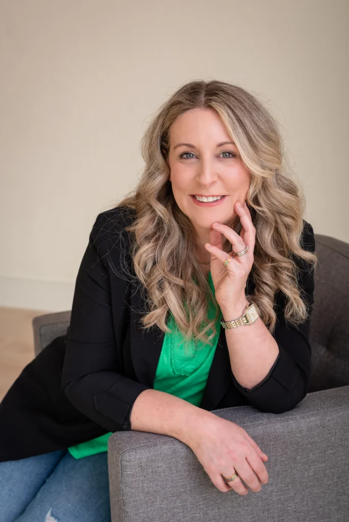 Smiling professional woman with long wavy blonde hair, wearing a green blouse and black blazer, seated on a grey armchair in a modern office setting.